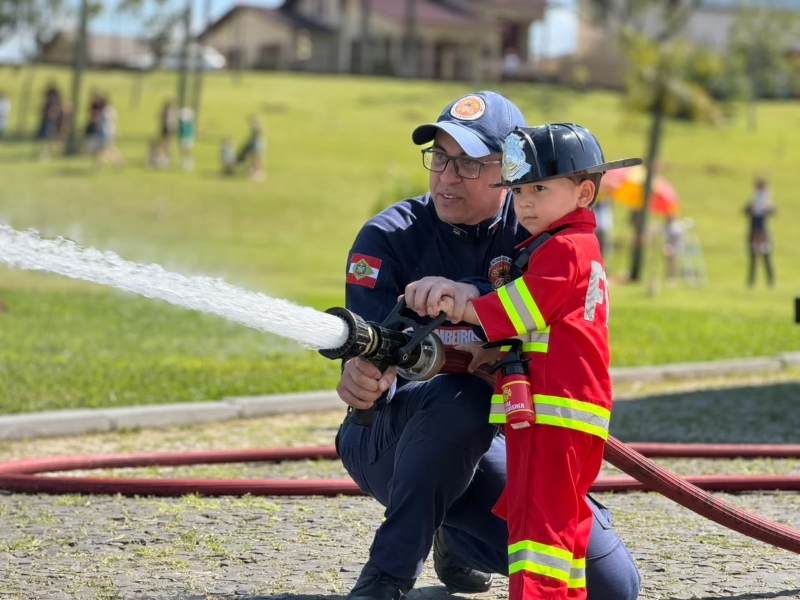 criança vestida de bombeiro segurando a mangueira com jato de água supervisionada por bombeiro