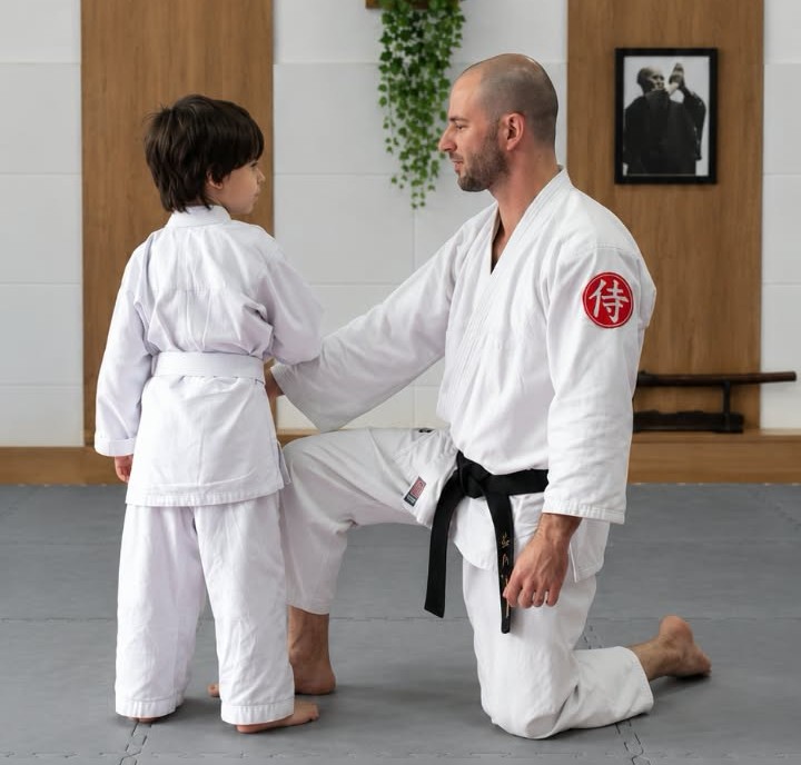 Sensei Willian Casagrande com um joelho apoiado no tatame, de frente para um aluno em pé. Ambos vestem quimonos brancos; o professor usa faixa preta e a criança, faixa branca