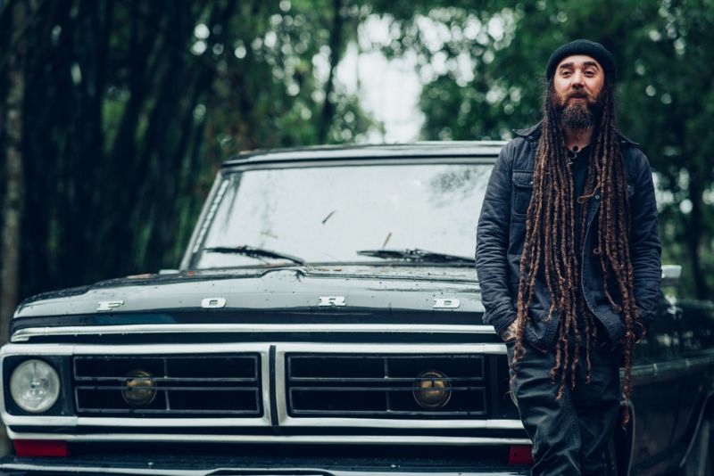 Victor Pradella, de gorro preto e cabelos longos com dreadlocks, posa diante de uma caminhonete Ford em uma área arborizada.